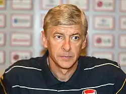 Photo serrée d'Arsène Wagner en conférence de presse avec les logos des sponsors floutés en fond.