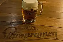 Une table en bois sur laquelle est posée une choppe de bière remplie de bière et de mousse ; la marque Staropramen est écrite à même la table.